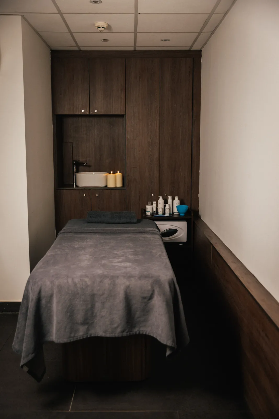 An empty treatment room with a made massage bed and wooden cabinets