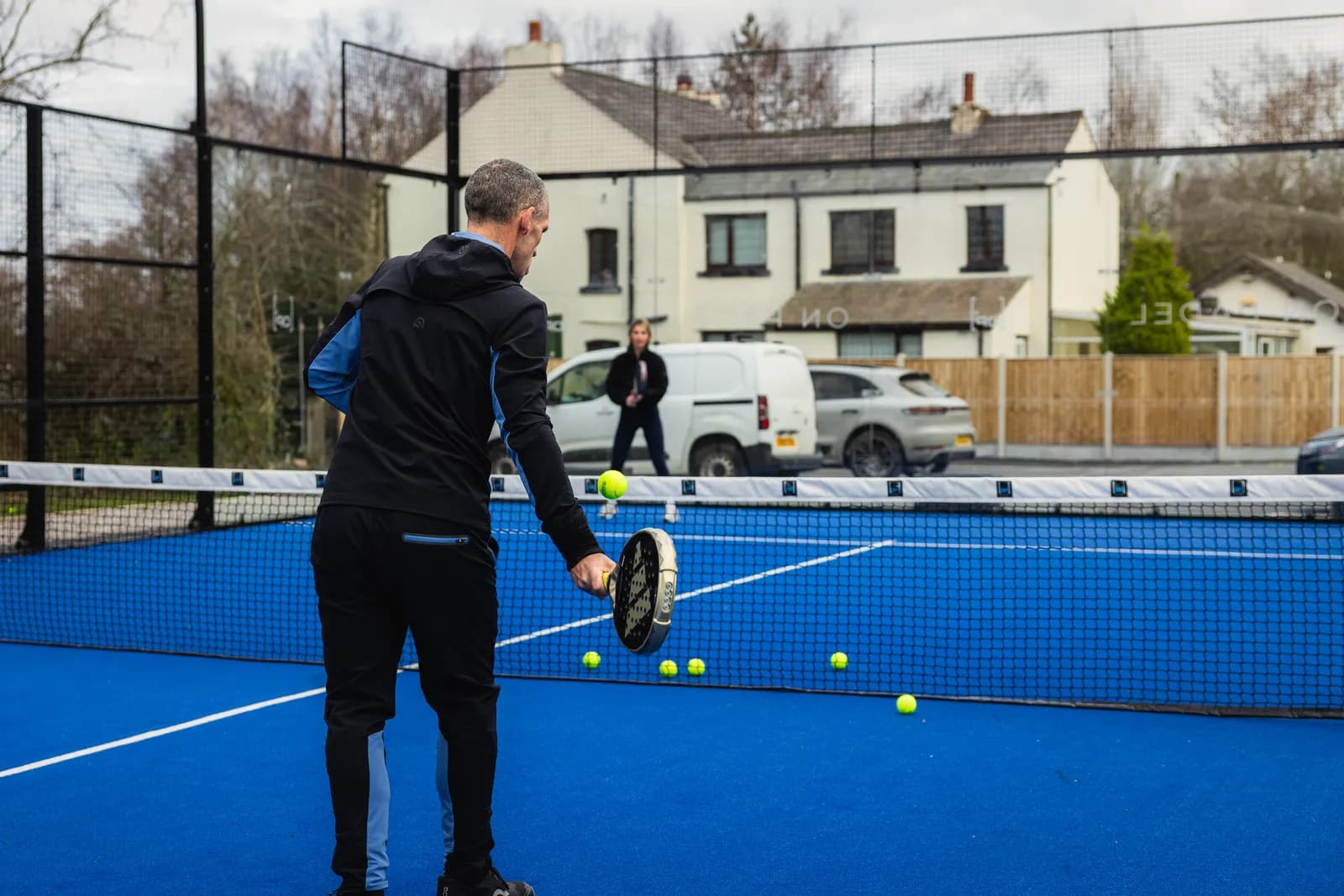 A coach feeding balls over the net on an outdoor padel court